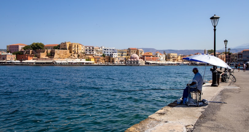 A person is sitting under a blue and white umbrella, fishing along a waterfront promenade. The background consists of a picturesque town with colorful historic buildings and a clear blue sky. Bicycles and street lamps line the path.